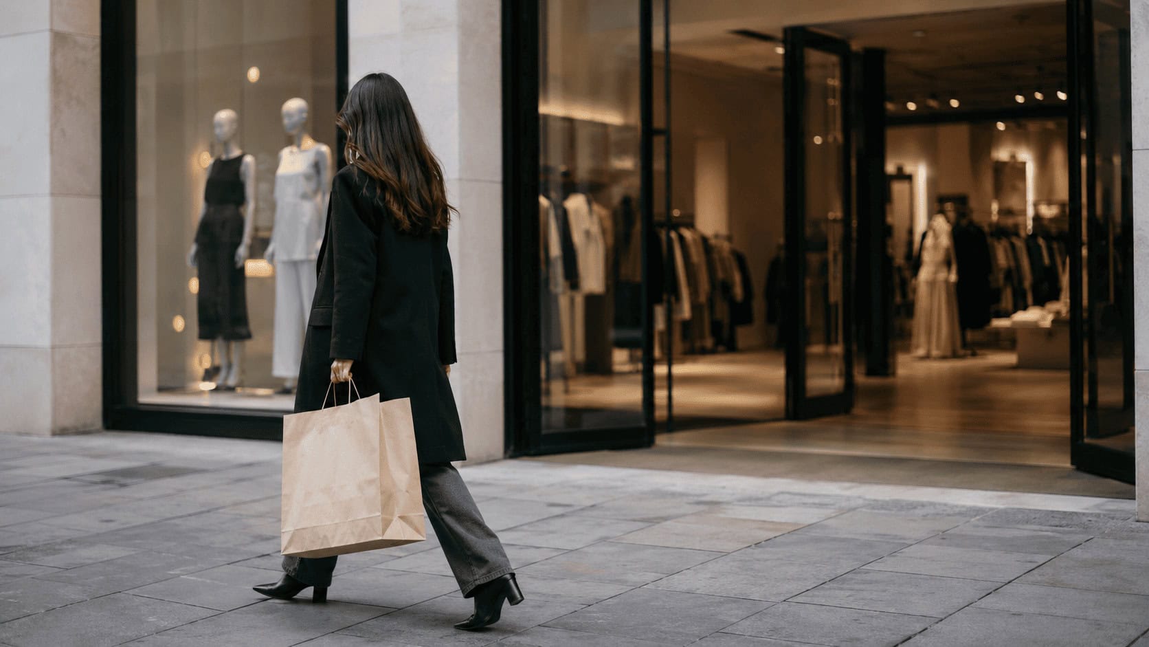 A shopper carrying a paper bag walks past the glass entrance of a Zara store on a city street in morning light.