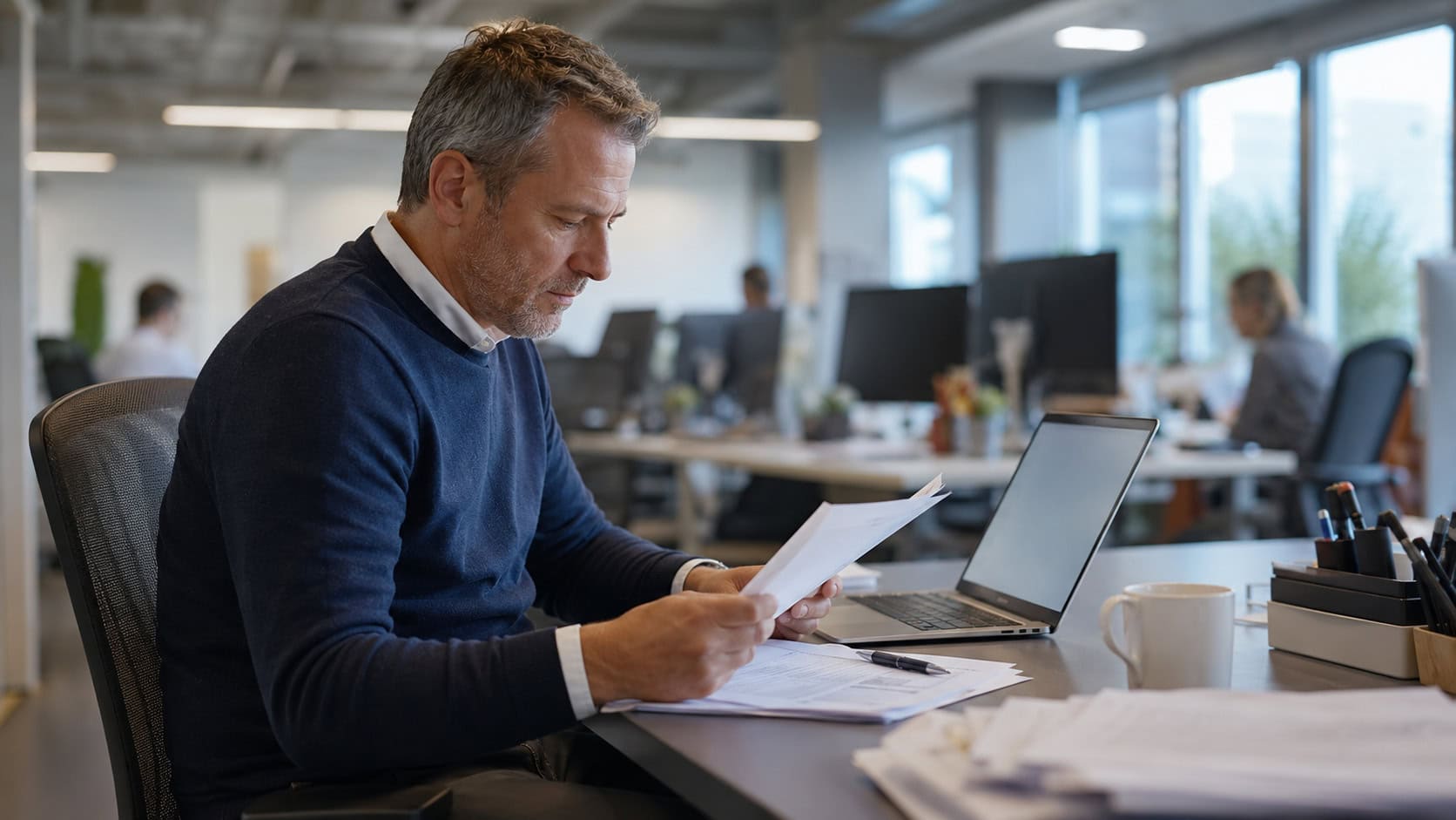 Manager reviewing printed documents beside a laptop in a modern open-plan office