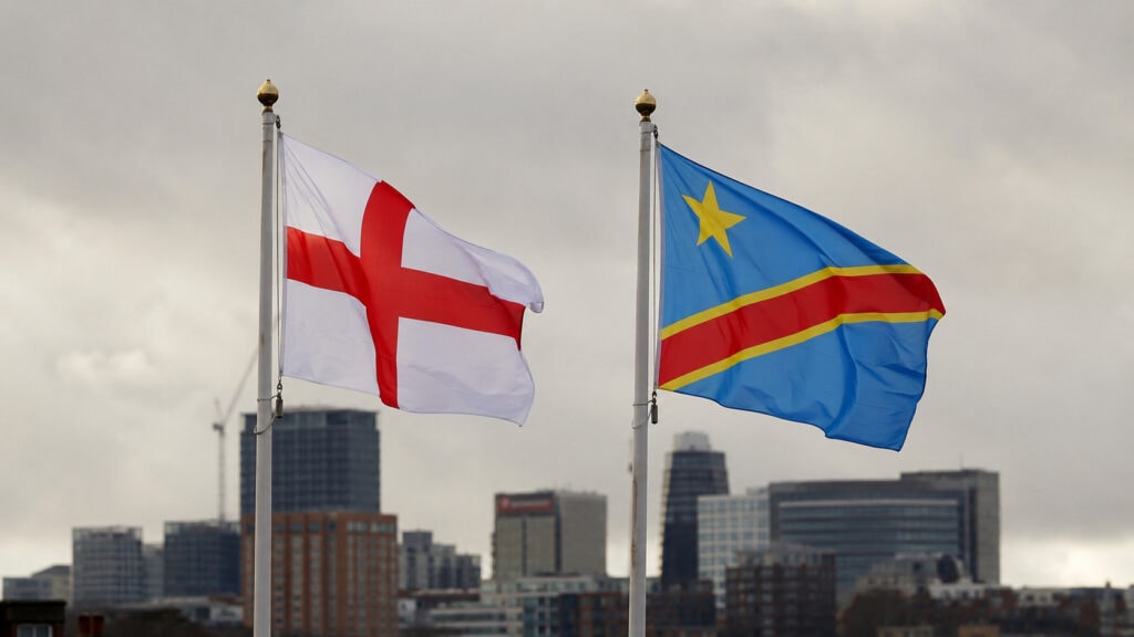 National flags of England and the Democratic Republic of Congo flying side by side against an overcast city skyline