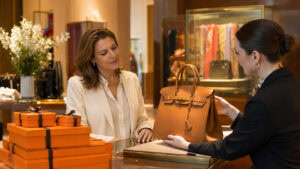 Affluent woman viewing a tan leather Birkin-style handbag with a sales associate in an Hermès boutique, illustrating the Hermès target market