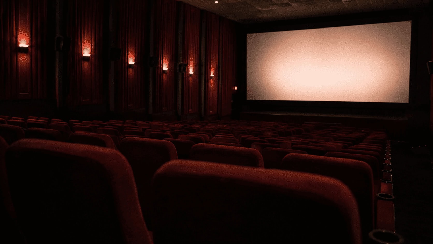 An empty movie theater auditorium lit in dim amber light with a blank glowing screen, evoking Hollywood's search for the next low-budget horror hit.