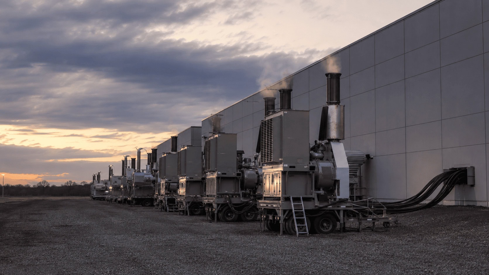 Row of trailer-mounted mobile gas turbines connected by cables to a large data center at dusk.
