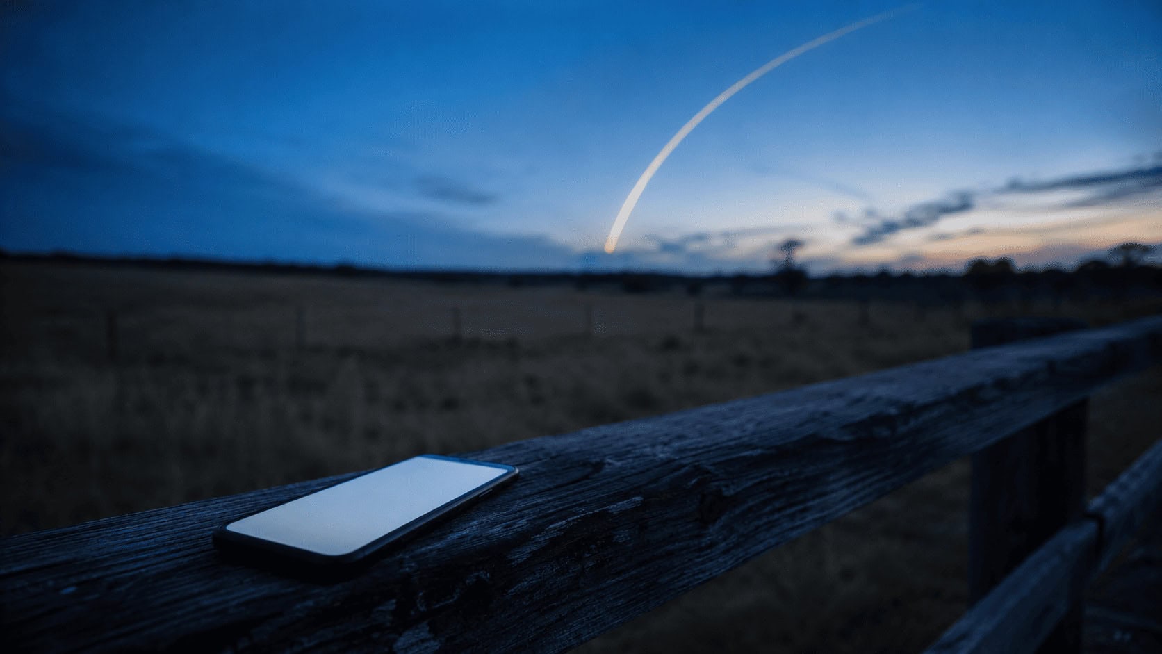 Starlink mobile concept: a smartphone glowing on a rural fence rail as a SpaceX rocket streaks across the twilight sky