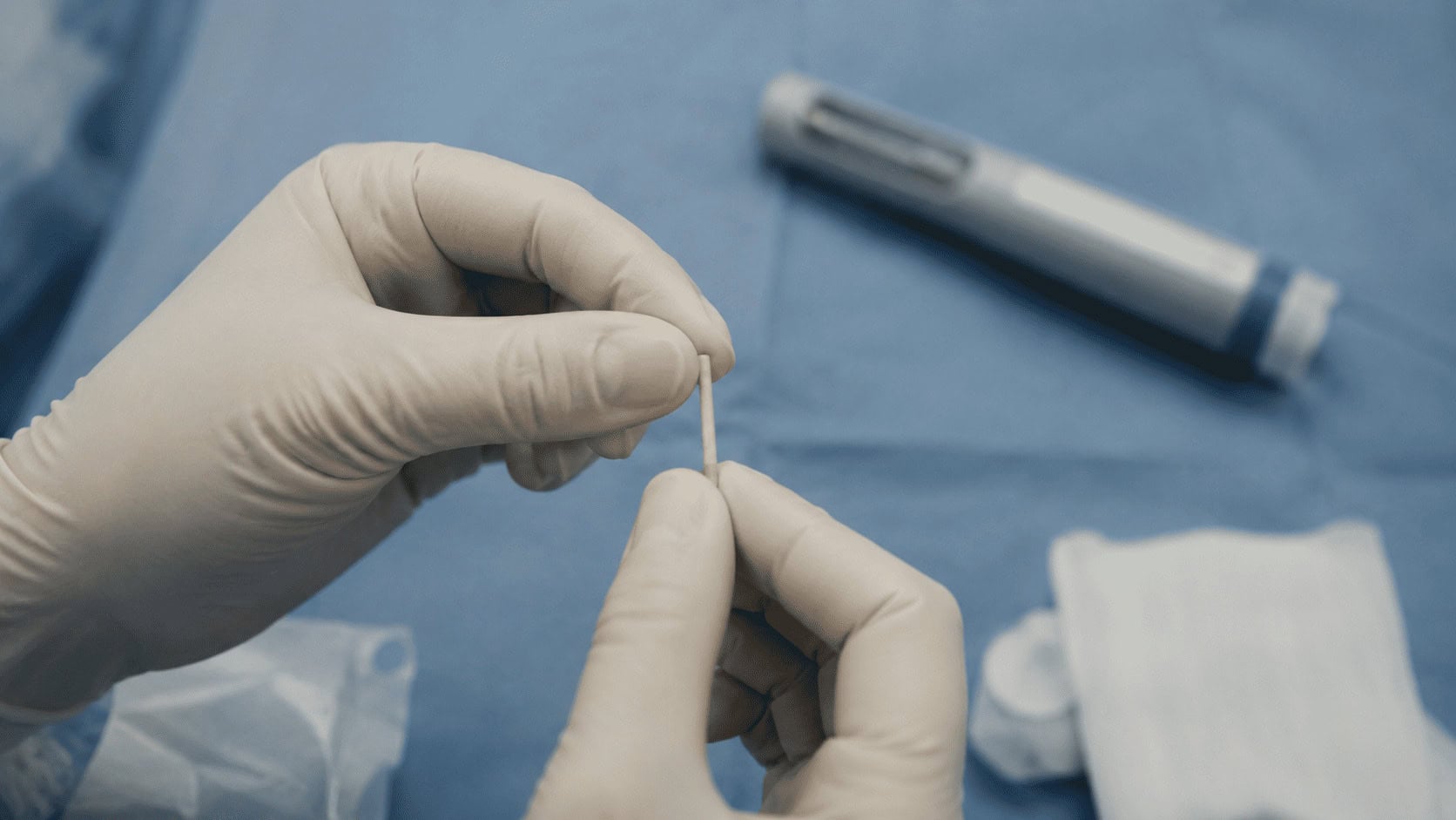 A matchstick-sized subdermal drug implant held in gloved hands, with a weekly GLP-1 injector pen blurred in the background.