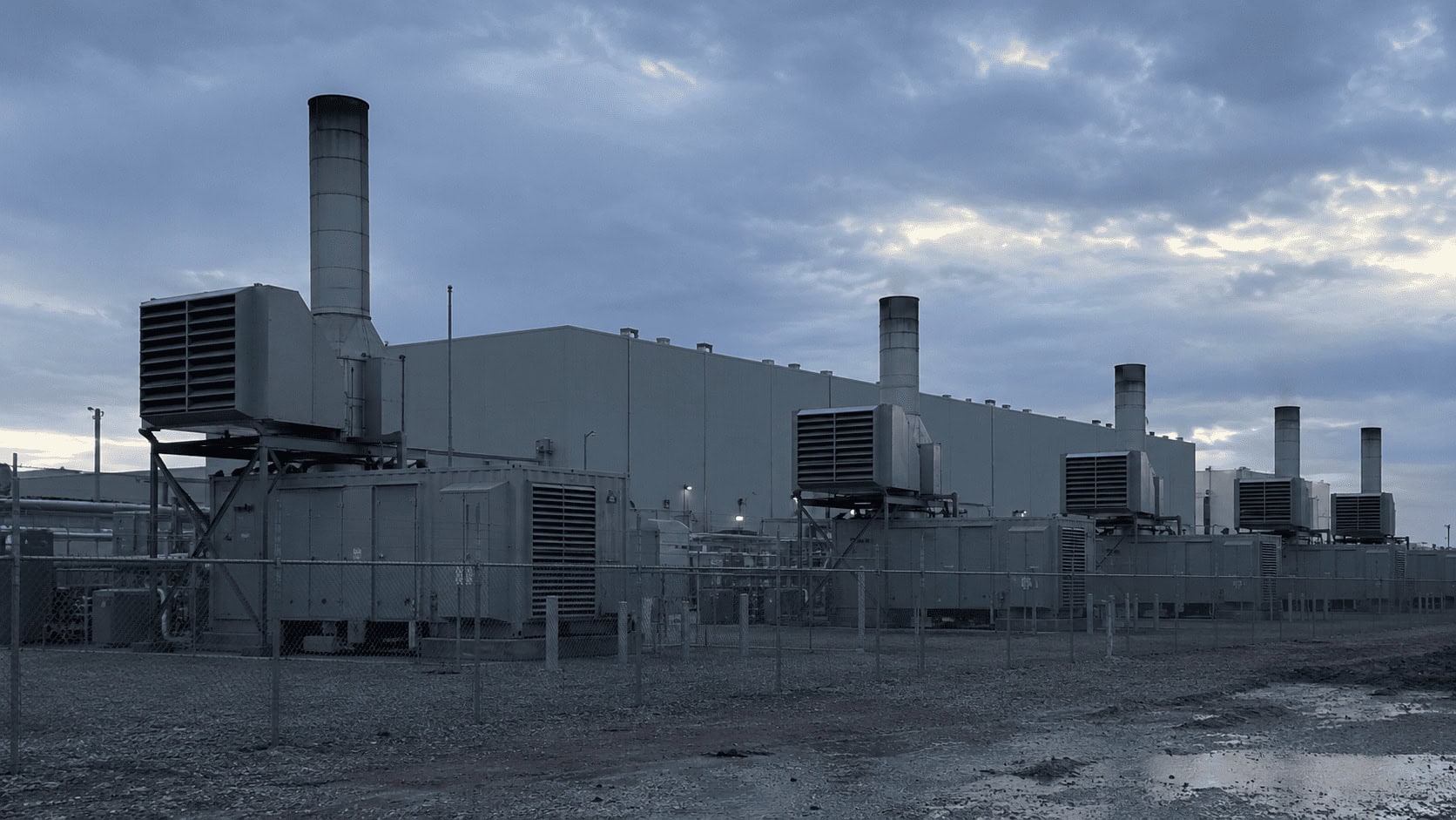 Modular gas turbine generators behind a fence next to a large data center building at dusk