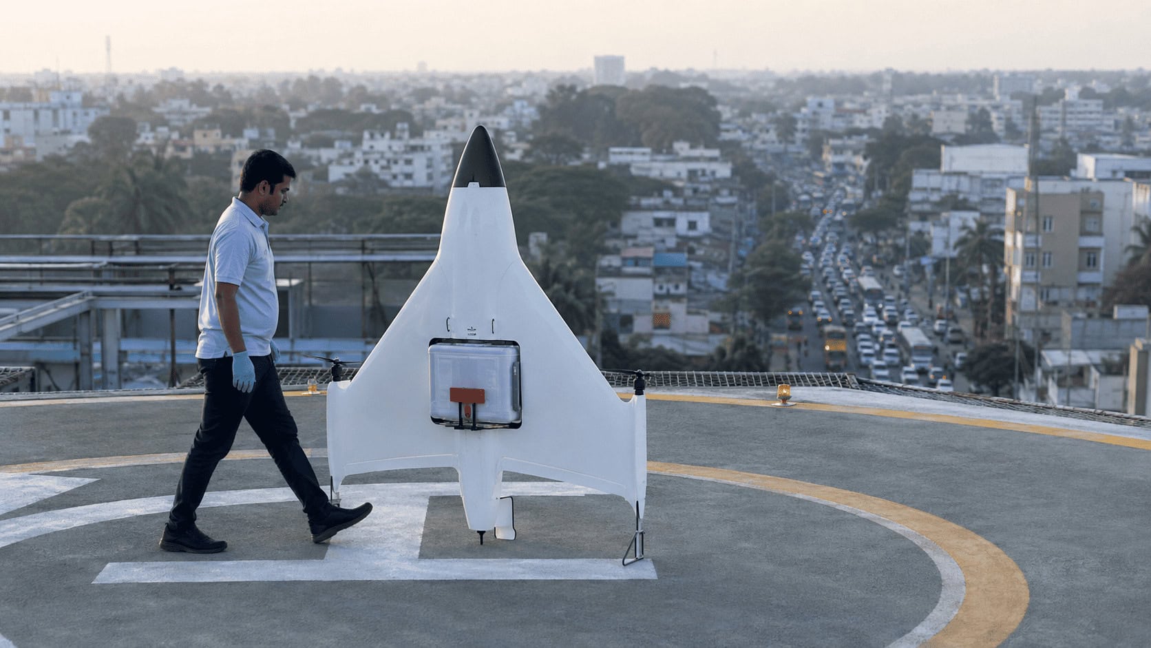 A carbon fibre tail-sitter delivery drone lifting off vertically from a hospital rooftop pad in Bengaluru with congested city traffic visible below