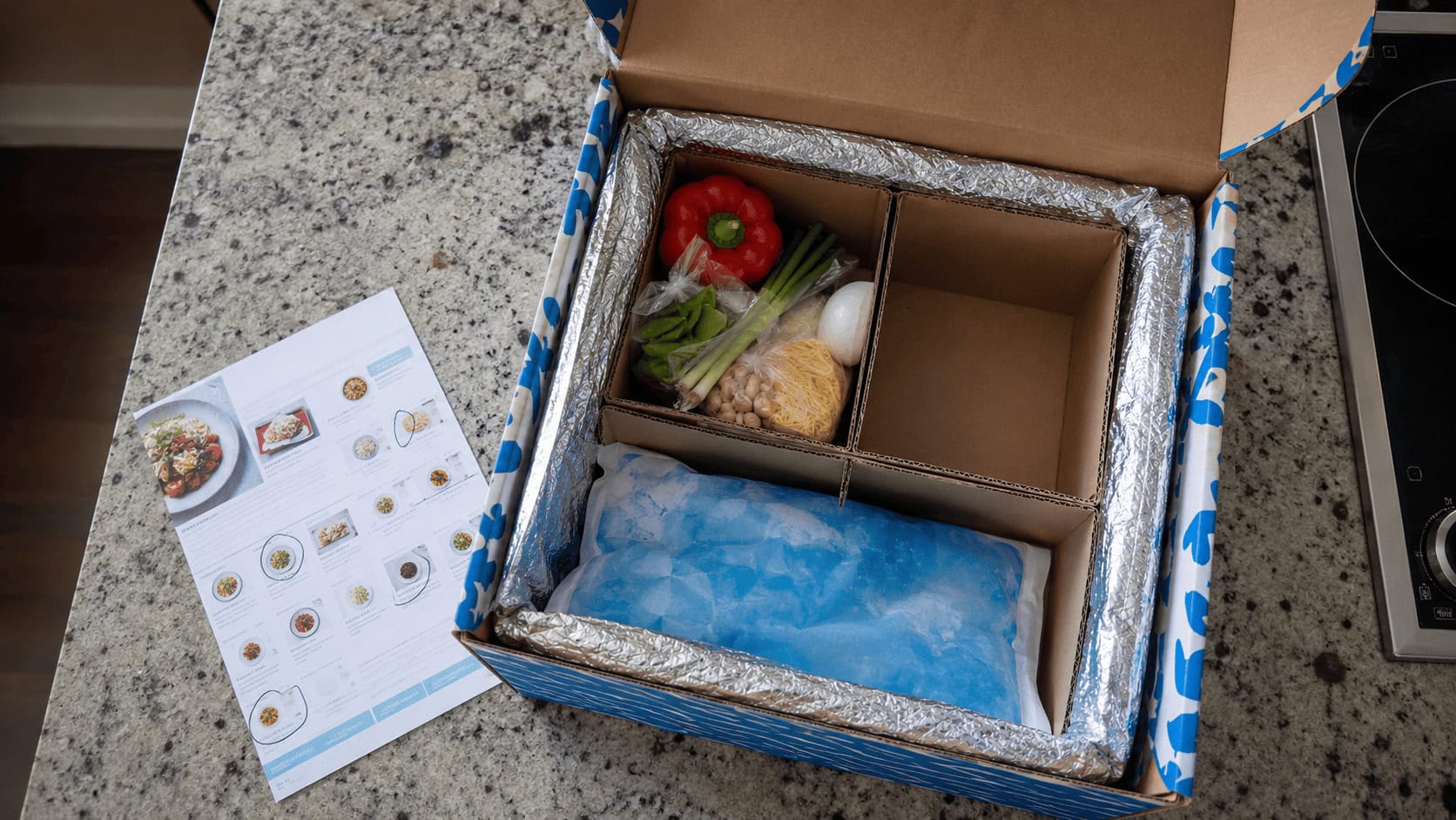 An opened Blue Apron style meal kit box on a kitchen counter with a recipe card and several missing ingredient slots