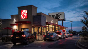 Servicio de autoservicio de Chick-fil-A al anochecer, con coches haciendo cola frente a un letrero rojo iluminado del restaurante y una valla publicitaria con la imagen de una vaca sobre el tejado