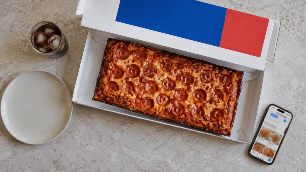 A rectangular single-serve pizza in an open Domino's-branded box on a kitchen counter beside a phone showing a food ordering app.