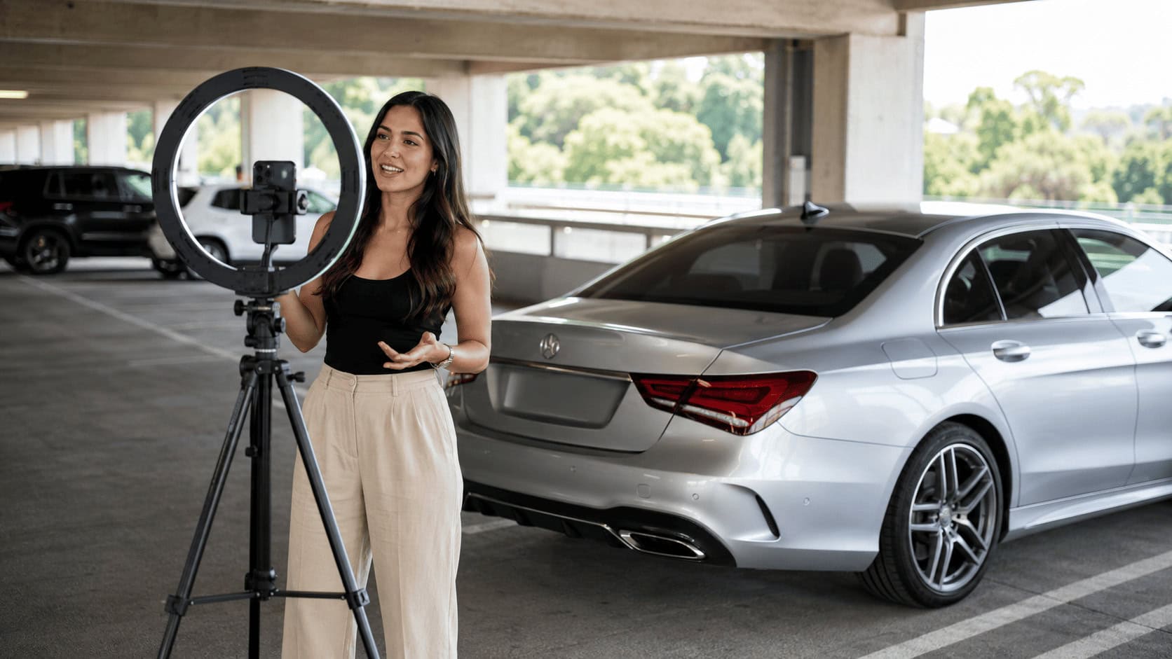 A young financial influencer films a smartphone video beside a silver luxury sedan in a parking garage, ring light on a tripod, phone mounted at eye level.
