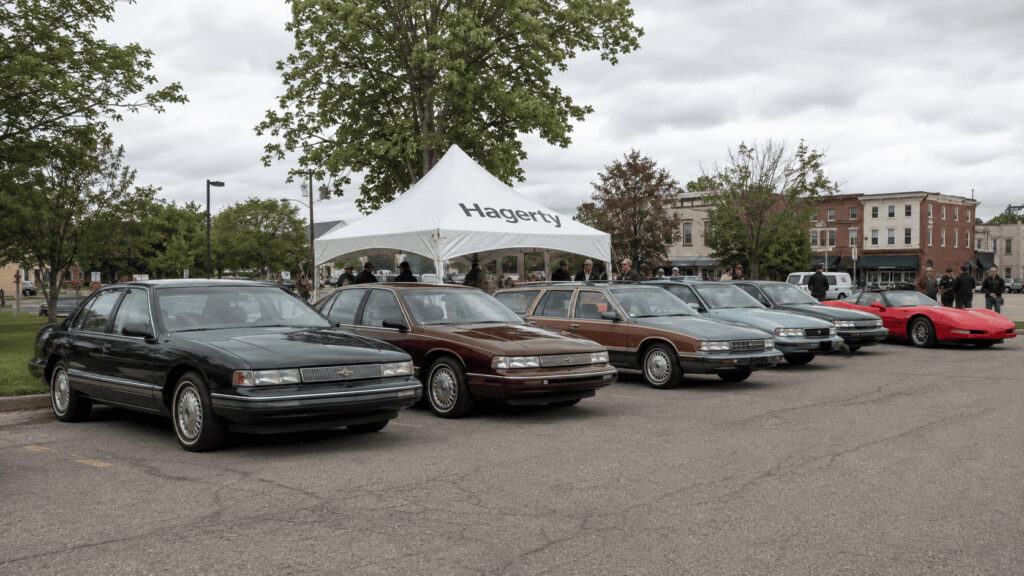 Row of 1990s American sedans and wagons parked at a suburban car show, with a Hagerty-branded event tent visible behind them