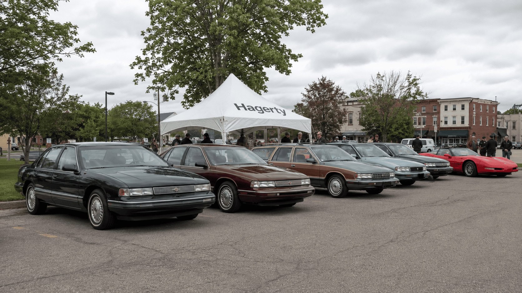 Row of 1990s American sedans and wagons parked at a suburban car show, with a Hagerty-branded event tent visible behind them