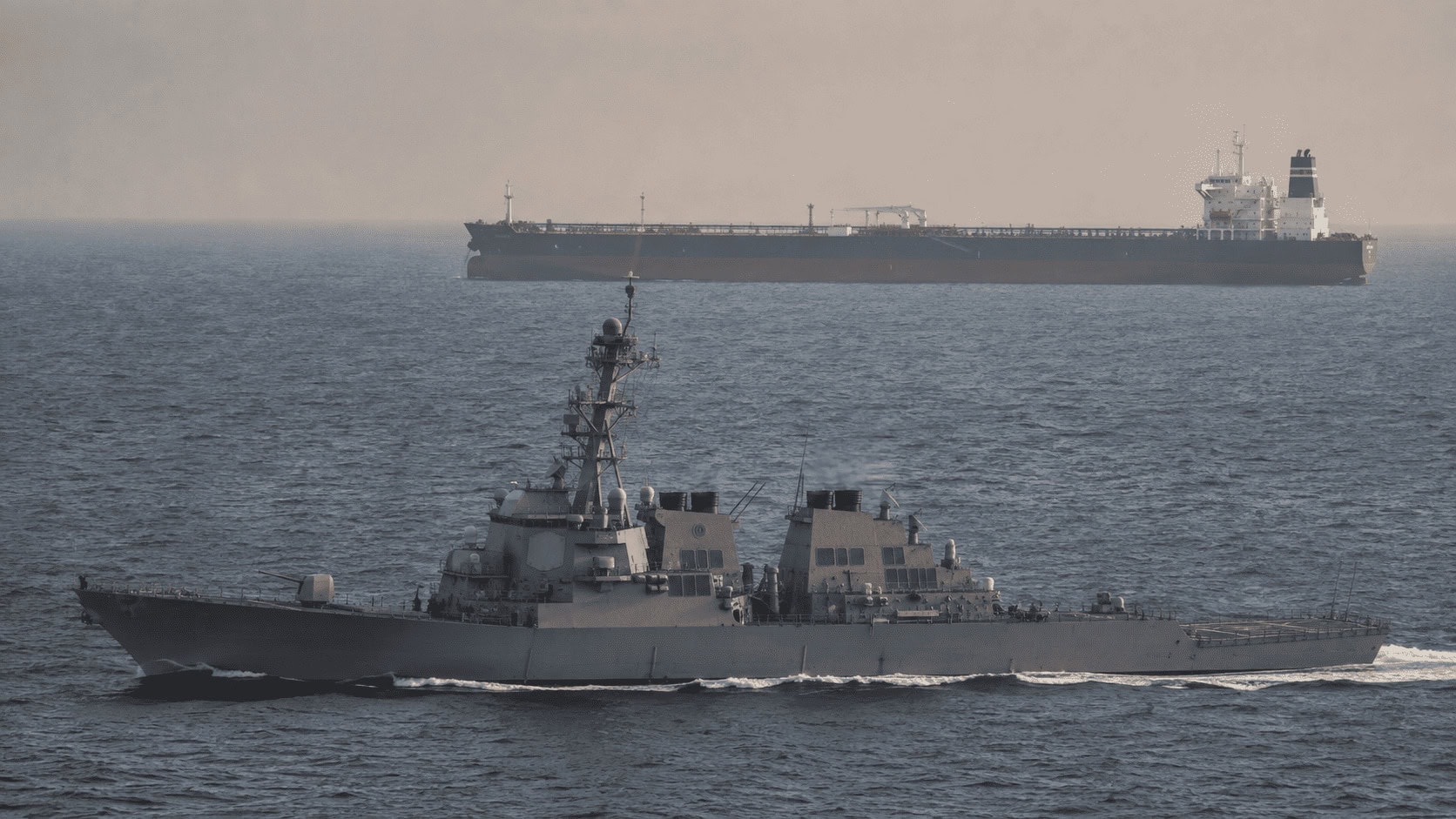A US Navy destroyer patrolling shipping lanes near an oil tanker in the Persian Gulf at dusk