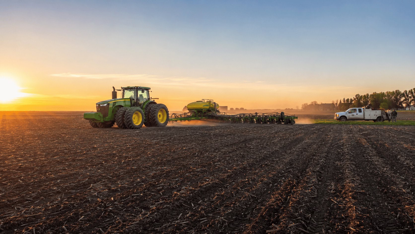 A John Deere tractor and planter working a Midwest field at sunset with a dealer service truck at the field edge.