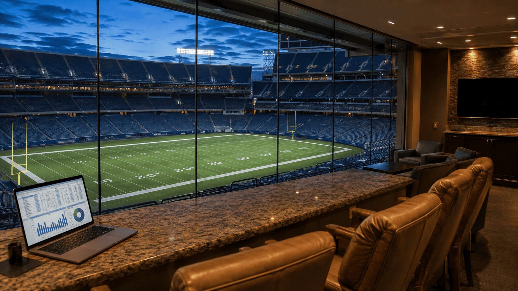 Empty luxury suite overlooking an NFL stadium field at dusk with a financial spreadsheet open on a laptop