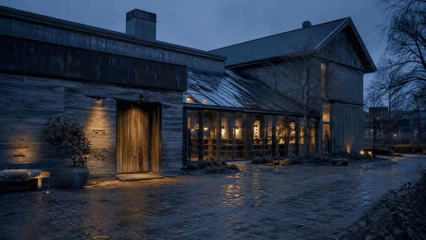 Exterior of the Noma restaurant on its Copenhagen campus at dusk, warm light glowing from greenhouse-style windows