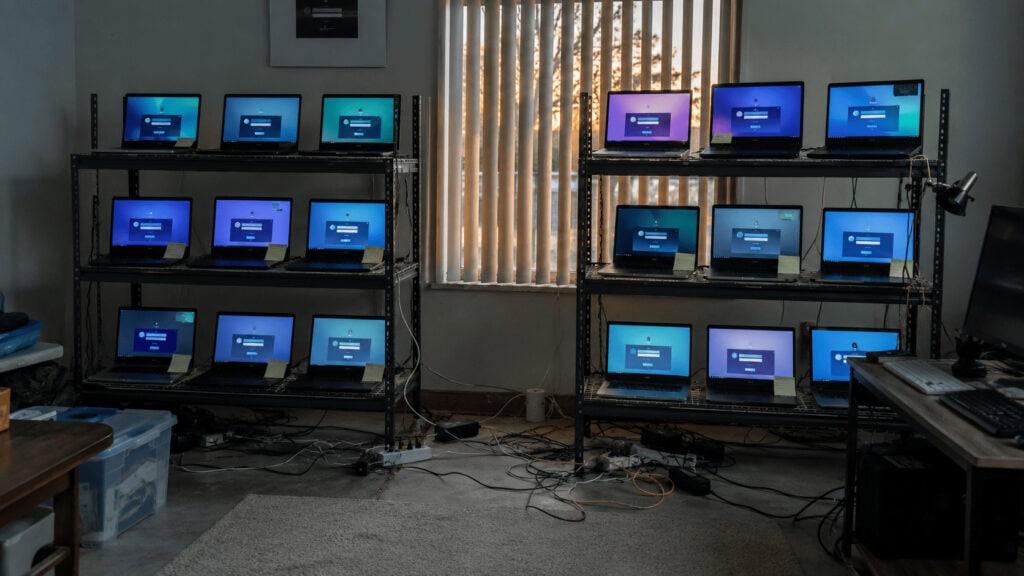 Rows of open corporate laptops on metal shelving in an American suburban home office, each screen showing an active remote desktop session