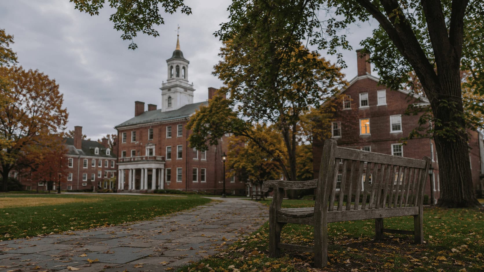 Empty brick quadrangle at a small New England private college on a late summer afternoon, with a closed admissions building in the background.