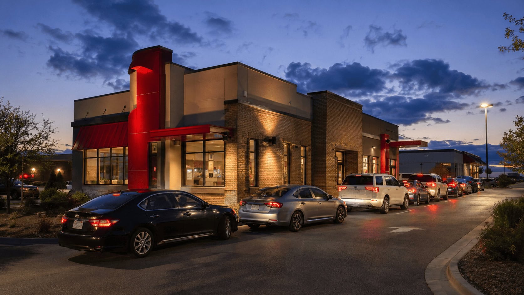 Raising Cane's Chicken Fingers drive-thru with the yellow and red logo lit at dusk and a line of cars queued at the window