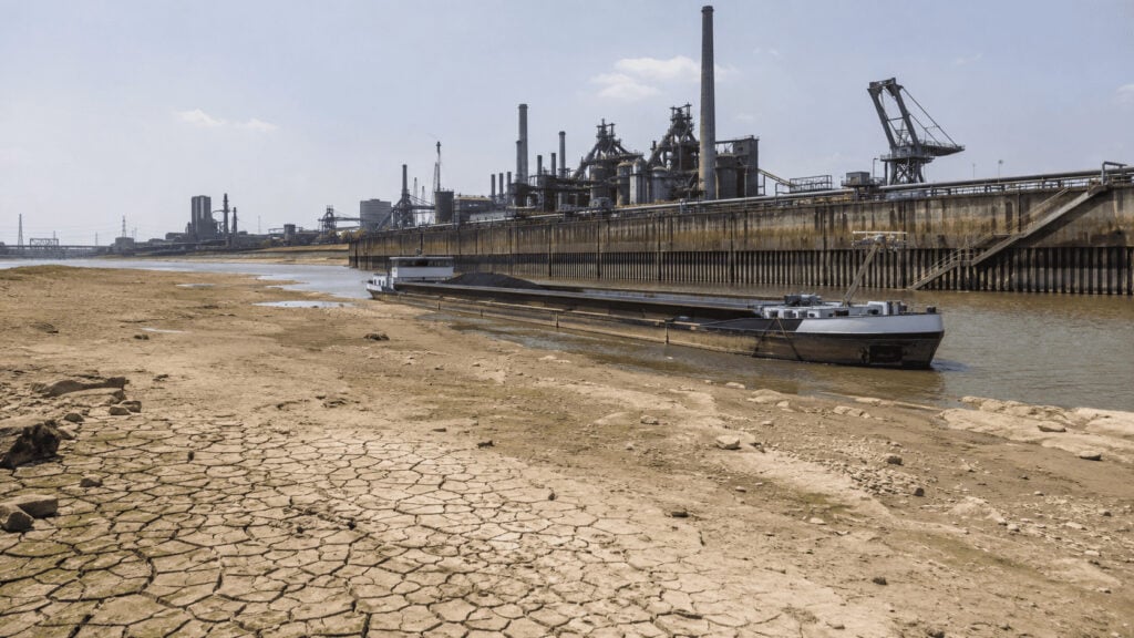 A cargo barge sits high in the water beside exposed Rhine riverbed at Duisburg, with ThyssenKrupp industrial works on the far bank