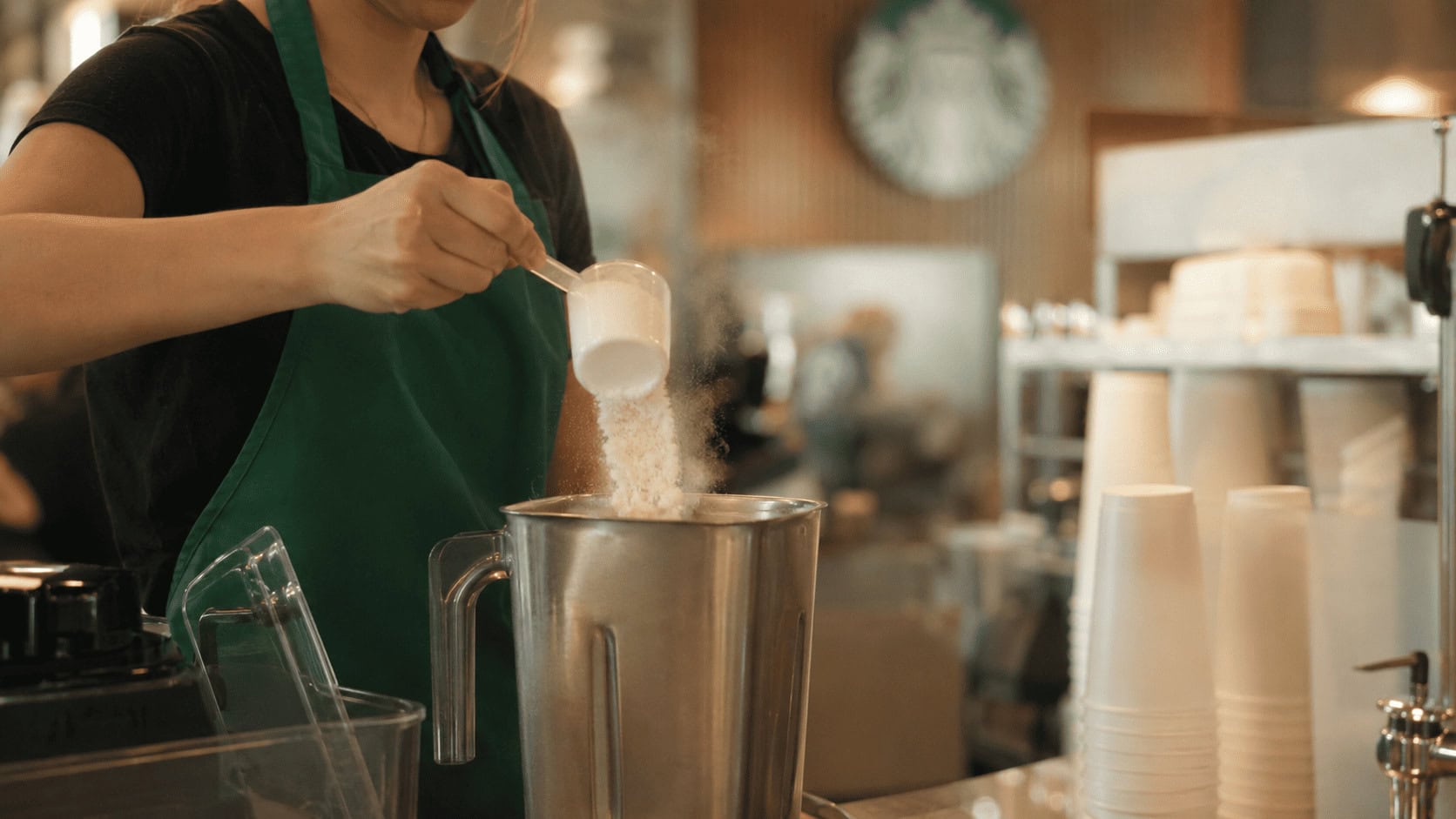 Barista in a green apron scooping fine white blending powder into a blender behind a Starbucks cold bar, a faint dust cloud catching the light