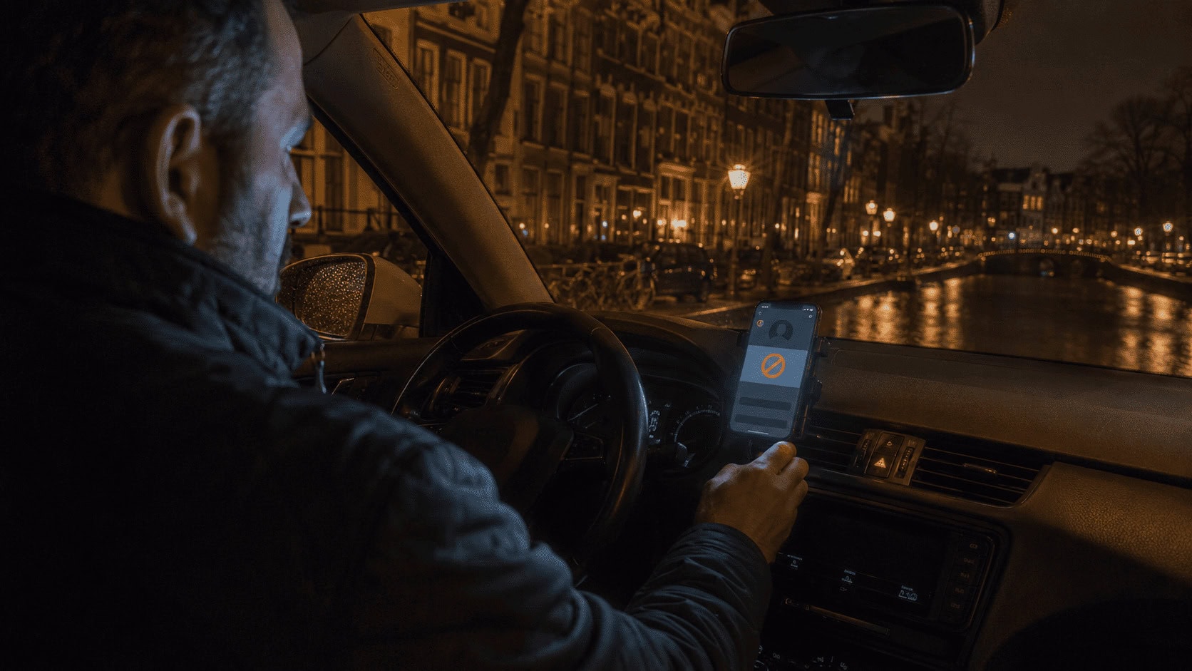 Ride-hail driver sitting in a parked car at night on an Amsterdam canal street, looking at a dash-mounted phone showing a deactivated driver account screen