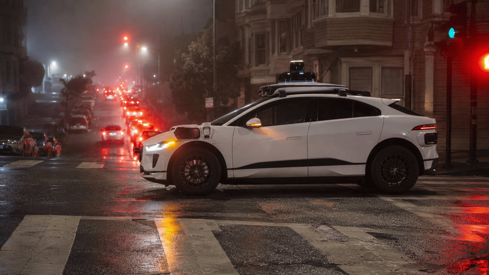 A white Waymo driverless Jaguar I-Pace with roof-mounted sensors stopped in heavy San Francisco night traffic, brake lights of blocked cars glowing behind it
