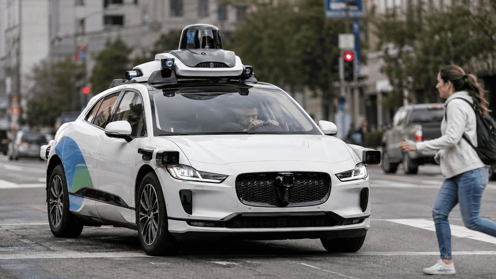 A Waymo autonomous Jaguar I-Pace with a human safety operator seated behind the wheel, driving a city street in daylight