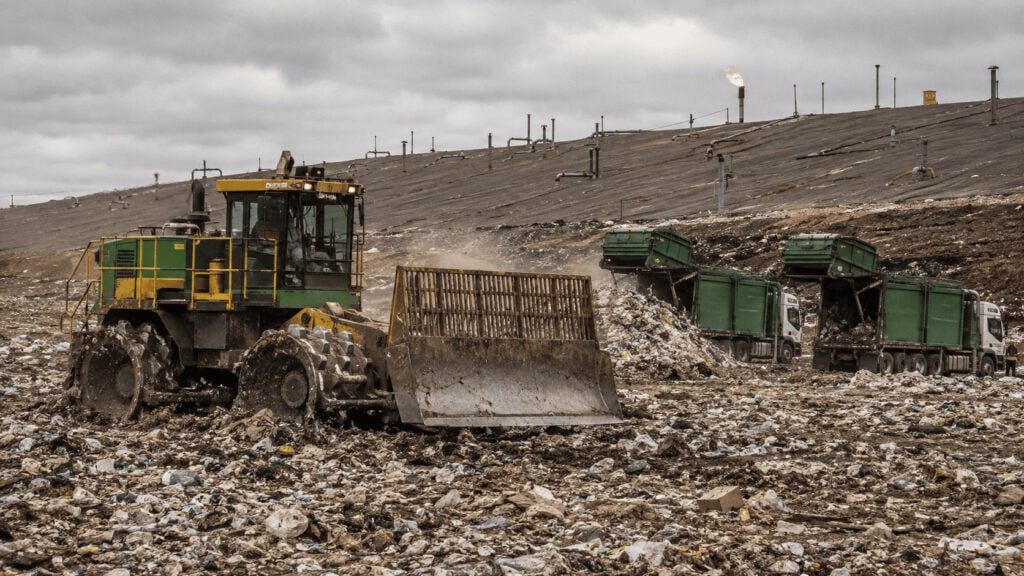 A WM landfill compactor working an active cell at a municipal solid waste site, with collection trucks queued at the tipping face