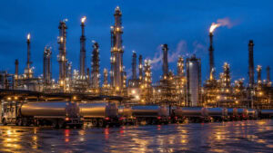 Tanker trucks lined up at a US refinery loading rack at dusk with distillation towers behind them