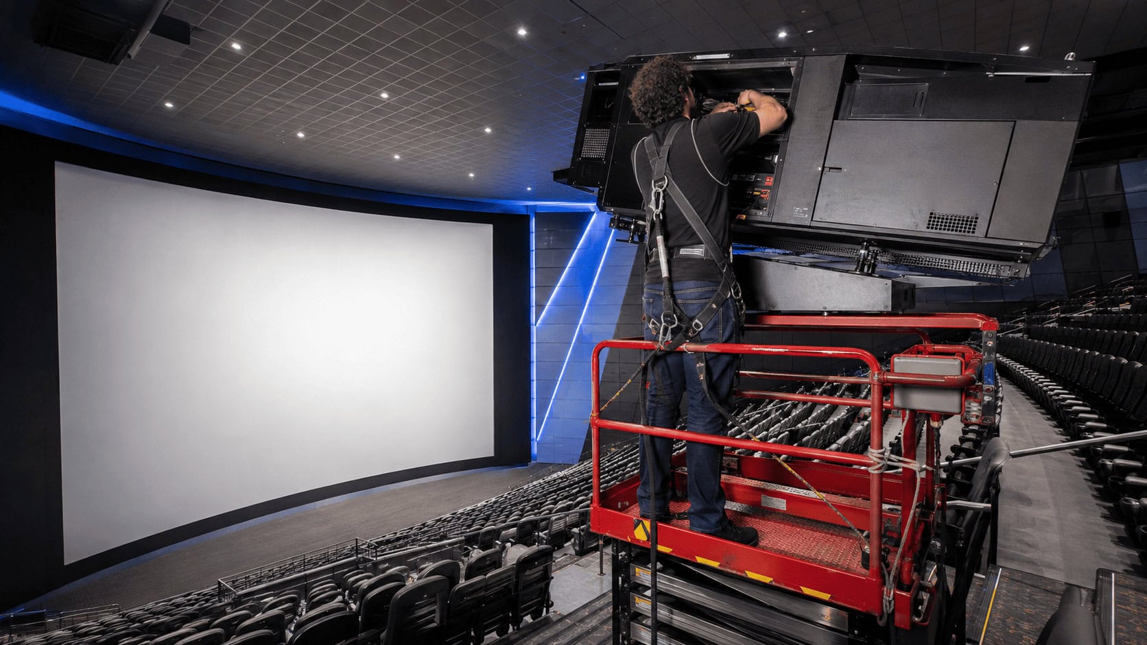 Technician on a scissor lift installing a large-format cinema projection system in an empty IMAX auditorium with the house lights on