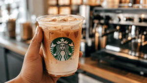 A hand holding a clear Starbucks cup of iced chai latte with the siren logo visible, condensation on the cup, on a store counter.