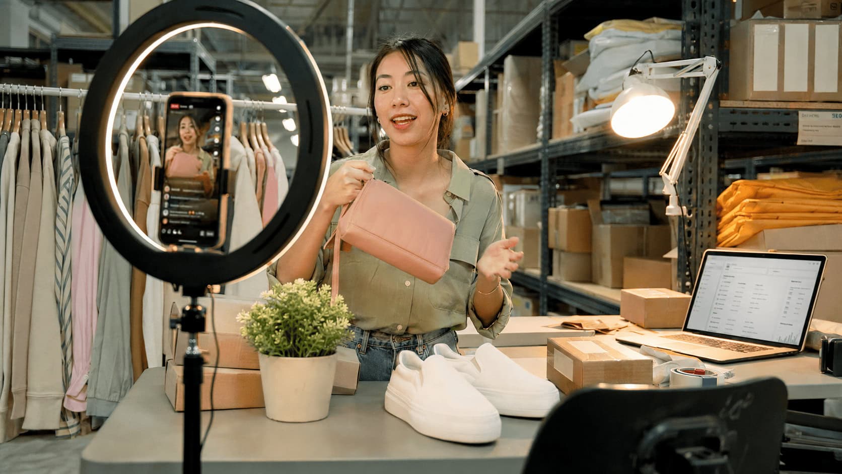 A livestream shopping host presenting beauty products to a ring-lit smartphone camera in a product-stacked warehouse studio, with a live viewer count and comment feed visible on the screen.