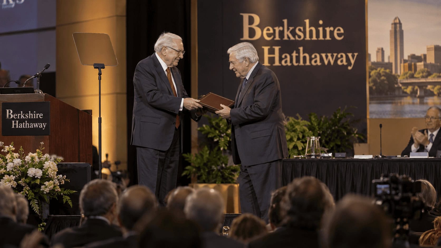 Warren Buffett sentado en una mesa de la reunión anual de Berkshire Hathaway mientras su hijo Howard Buffett toma el asiento del presidente a su lado