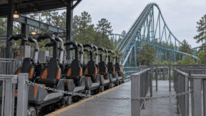 Empty roller coaster train parked with lap bars raised at a closed theme park station, with a tall steel coaster lift hill behind it.